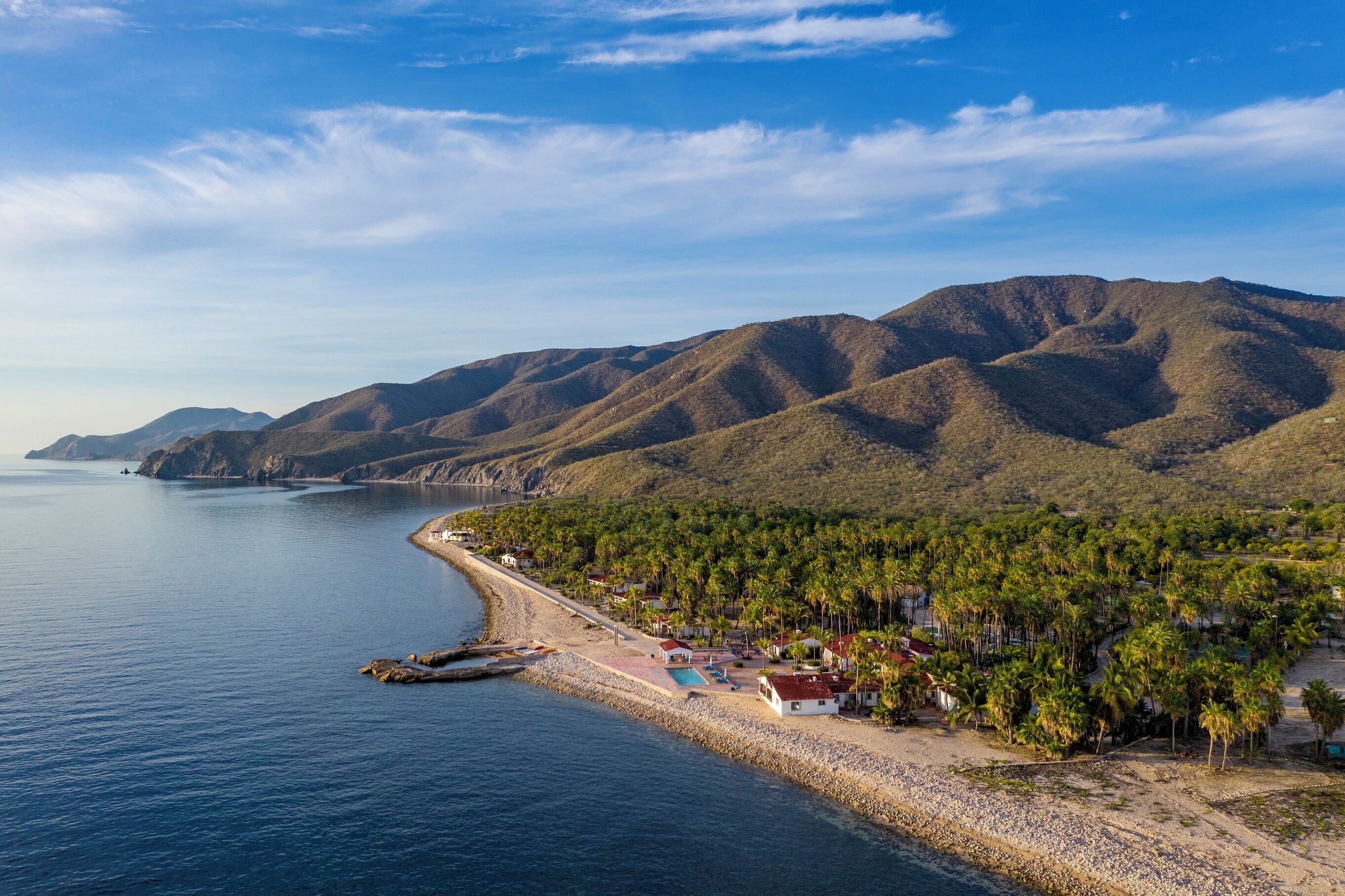 Un resort costero con playa, palmeras y montañas al fondo.