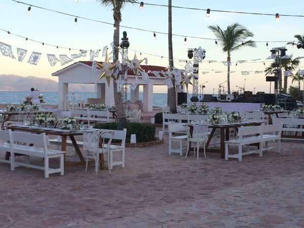 An outdoor wedding setup with white tables and chairs, a stage with musical instruments, and a backdrop of palm trees and mountains. - Rancho Las Cruces Baja California Sur (Las Cruces)