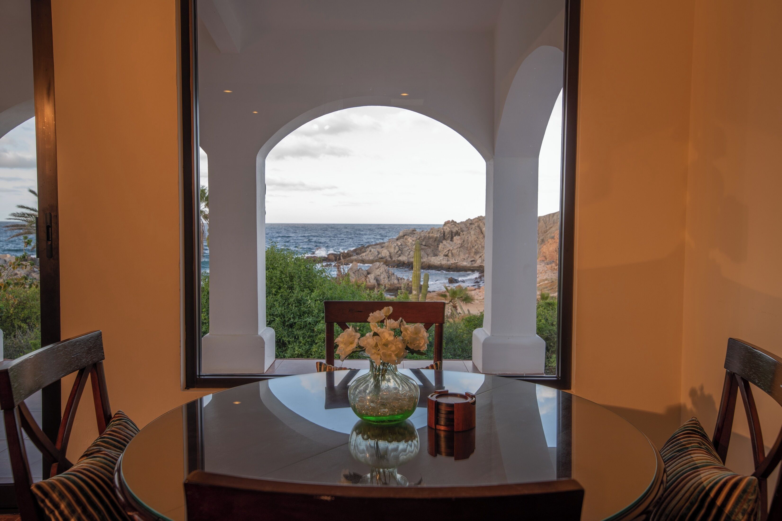 Une table à manger avec des chaises, un vase rempli de fleurs et une vue sur l’océan par une fenêtre en arc de cercle.