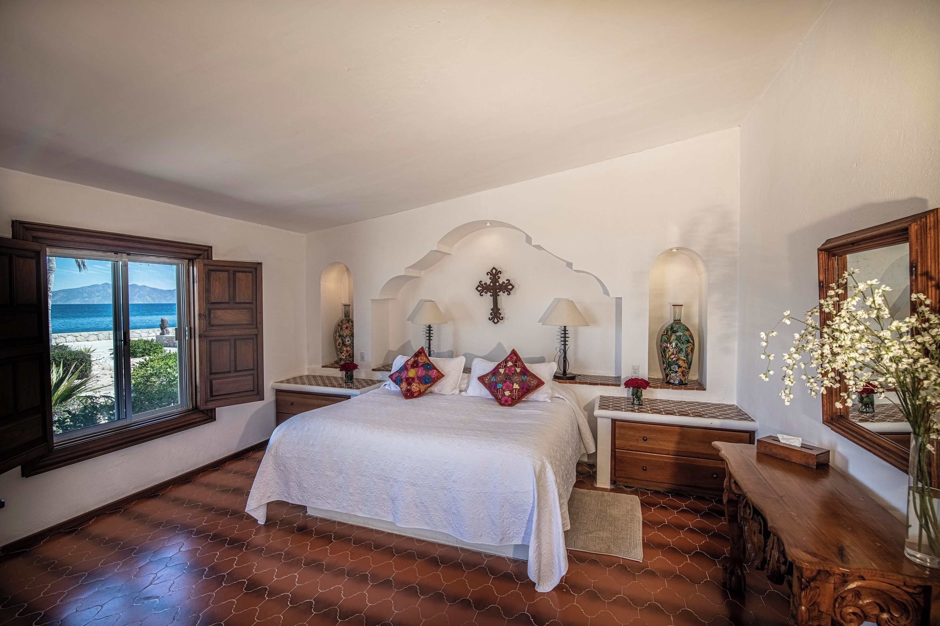 A bedroom with a large bed, a view of the sea through a window, and a decorative wall with a cross.