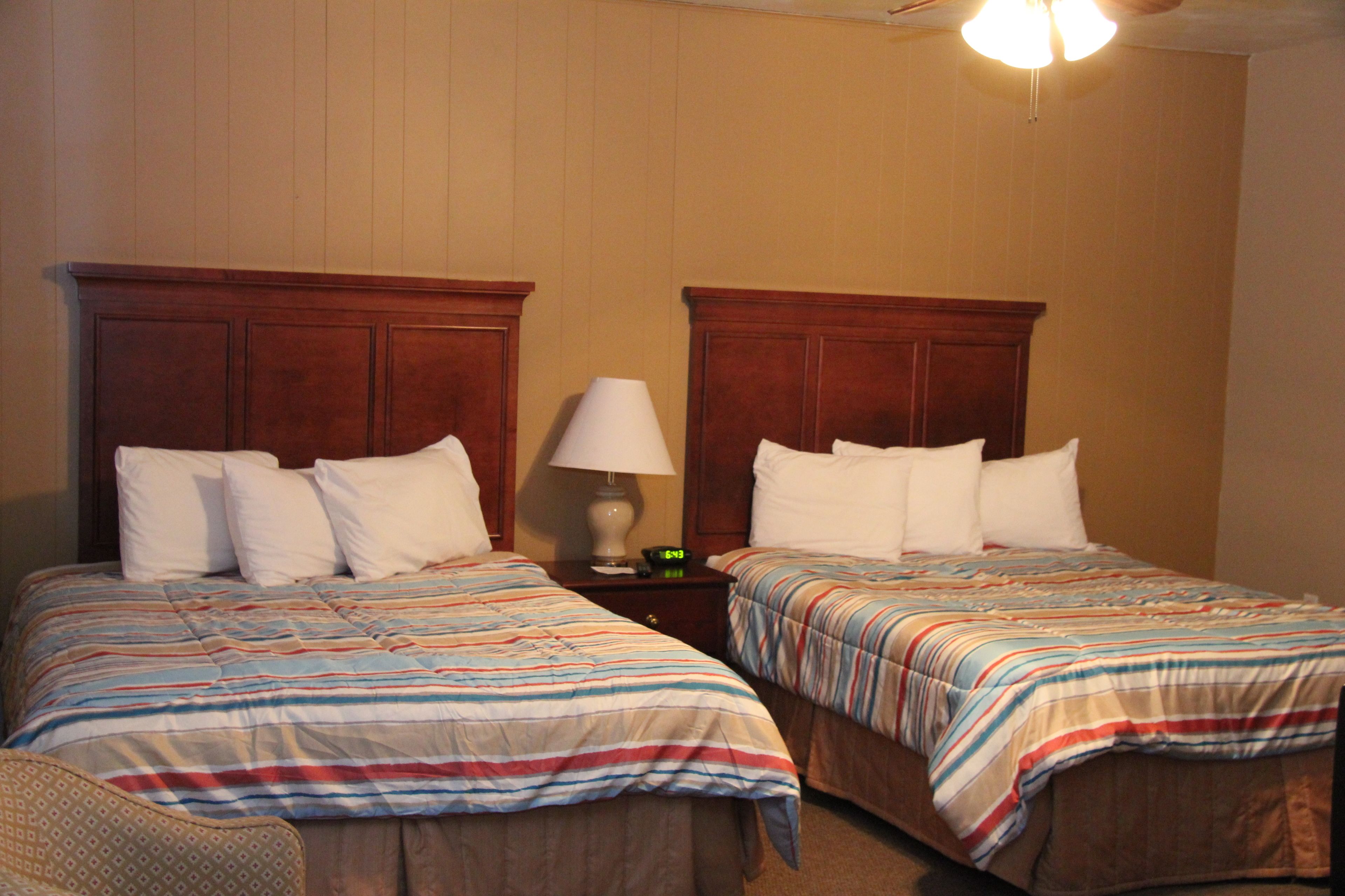 A hotel room with two single beds, wooden headboards, a nightstand with a lamp, and a small table.