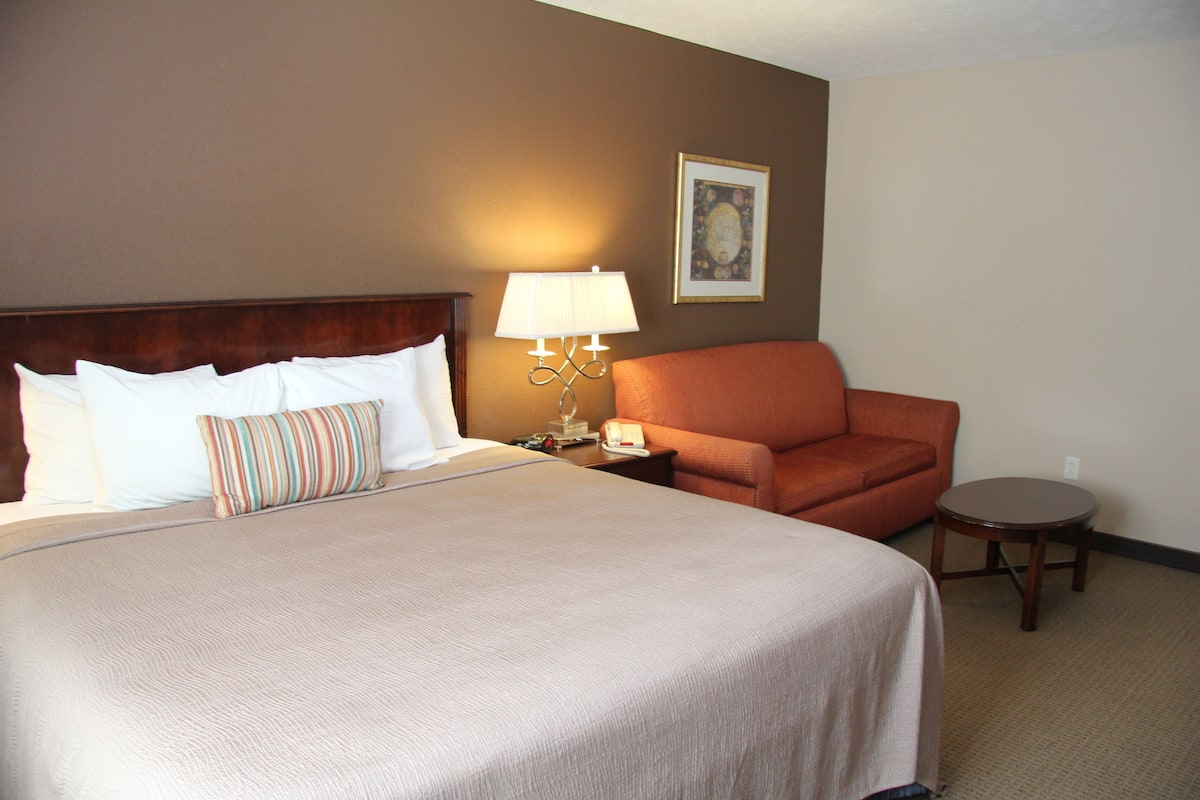 A hotel room with a large bed, a chair, a small table, and a lamp.