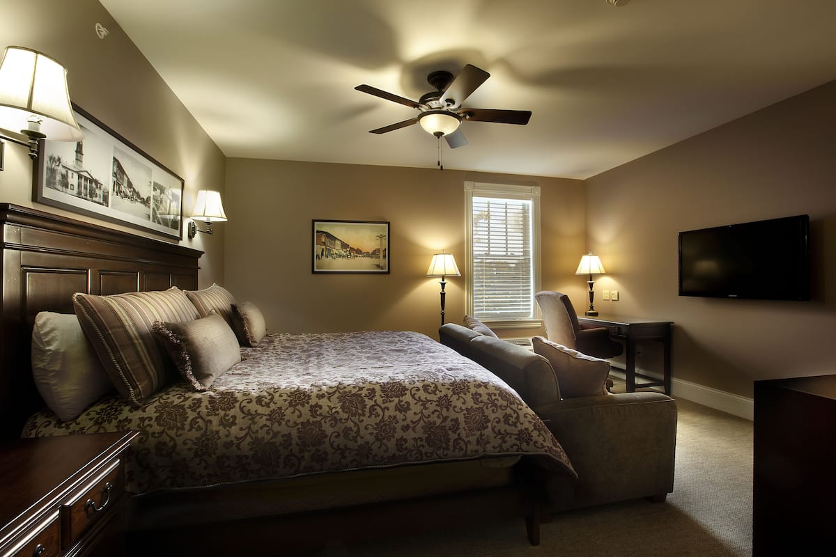 A bedroom with a bed, a chair, a desk, a ceiling fan, and a TV.
