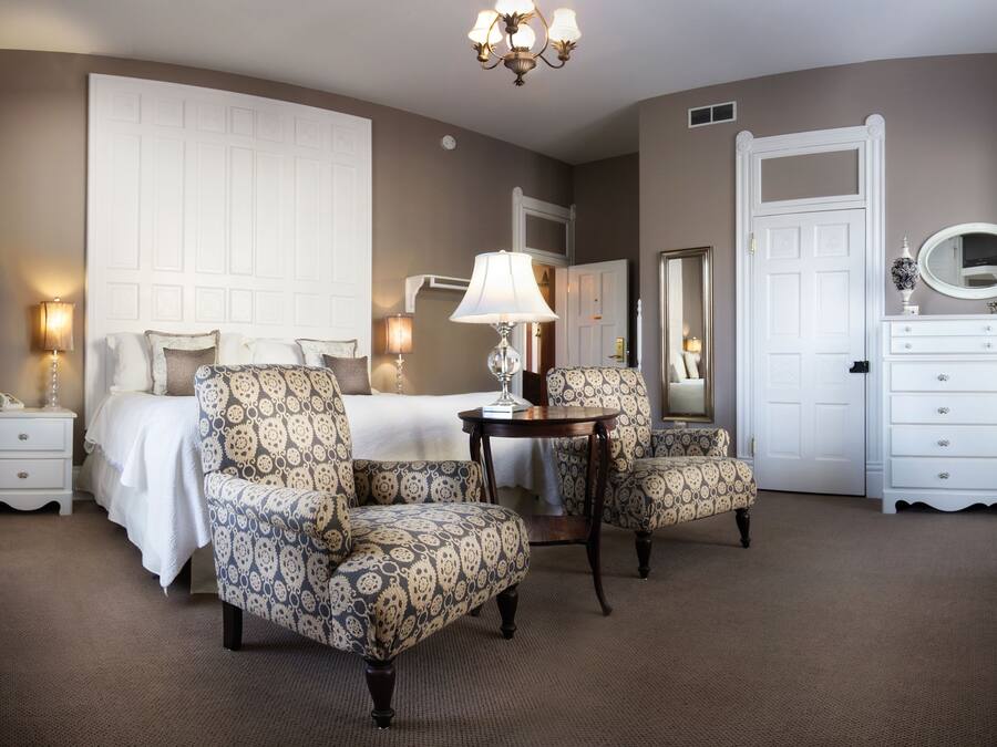 Une chambre d’hôtel comprenant un lit, deux fauteuils à motifs, une petite table avec une lampe, une commode et un miroir.