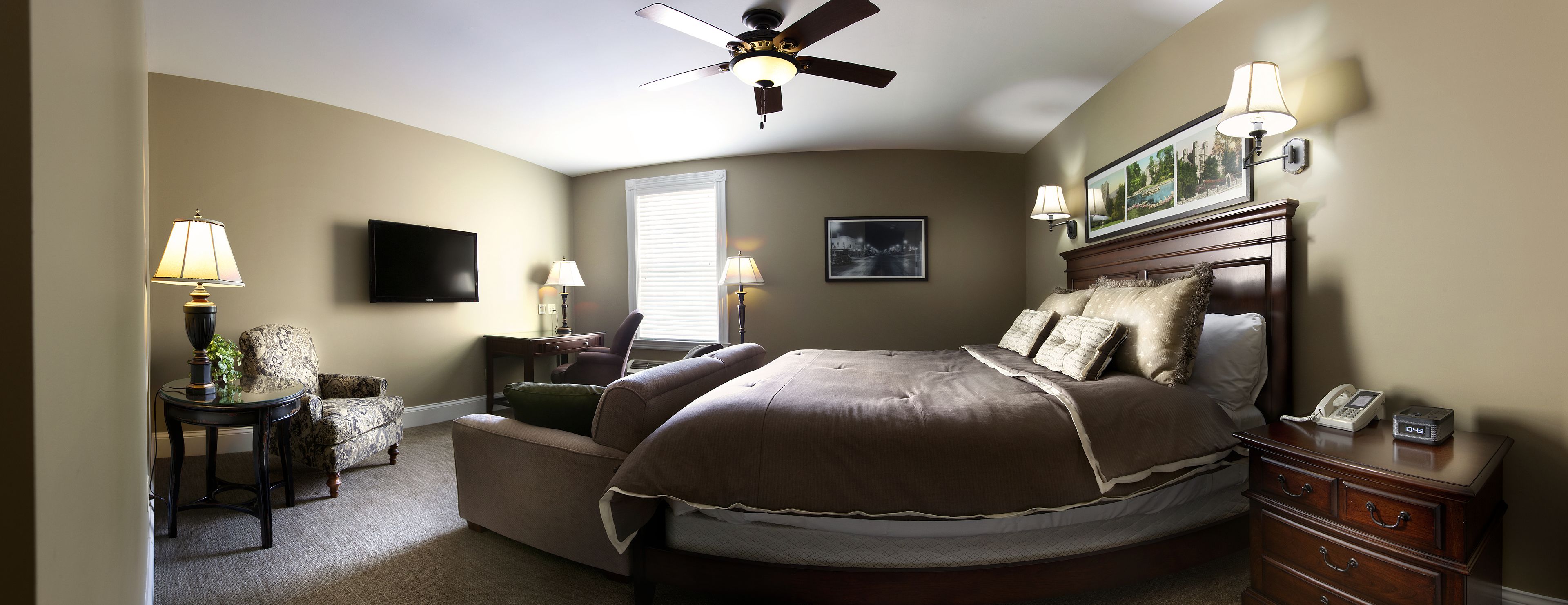 A bedroom with a bed, a desk, a chair, a ceiling fan, and a TV.