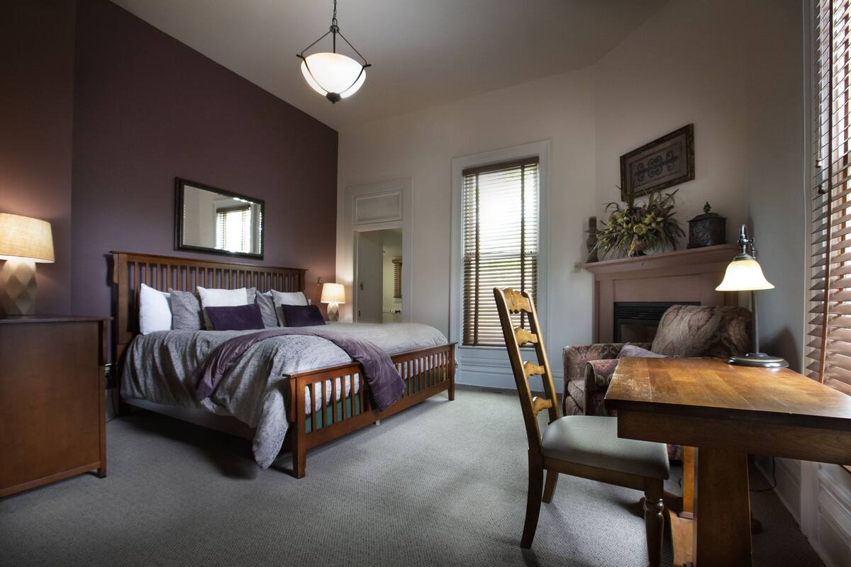 A bedroom with a bed, bedside table, chair, lamp, and a fireplace.