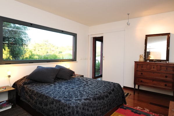 Une chambre avec une grande fenêtre, un lit recouvert d’une couvre-lit à motifs, une commode et un miroir.