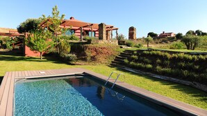 A swimming pool with a wooden deck, surrounded by a landscaped garden and a large house with a red roof. - Eco Suites Resort (Santiago do Cacém)