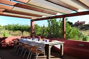 Une terrasse extérieure avec une table dressée pour le dîner, entourée de chaises en osier, et donnant sur une colline verdoyante avec un bâtiment en arrière-plan.