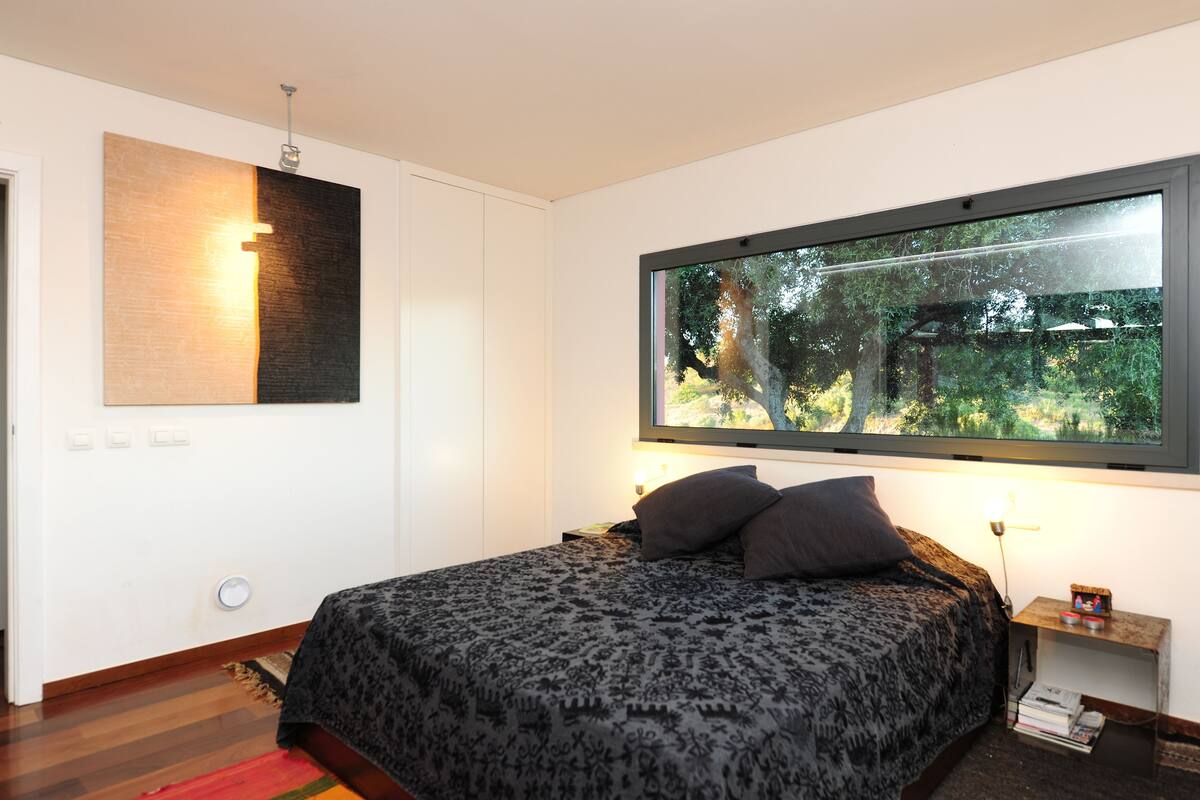 A bedroom with a large window, a bed with patterned bedding, a nightstand with a lamp, and a wall-mounted light fixture.