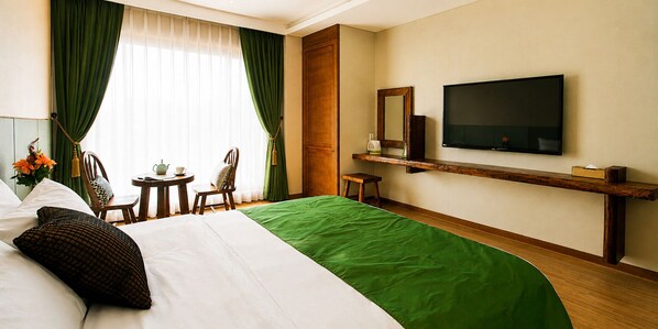 A hotel room with a bed, a TV, a small table, chairs, and a window with curtains.