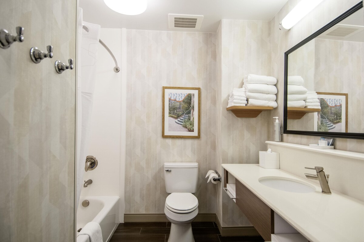 A bathroom with a bathtub, toilet, sink, and a mirror. There are towels stacked on a shelf and a framed picture on the wall.