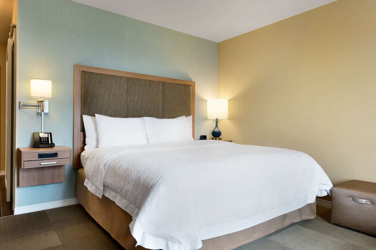 A hotel room with a large bed, a bedside table with a lamp, a telephone, and a small drawer.