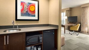 A hotel room with a kitchenette, a microwave, a refrigerator, a sink, a framed floral artwork, a television, a desk, a chair, and a window with curtains. - Hampton Inn & Suites Monroe (Monroe)