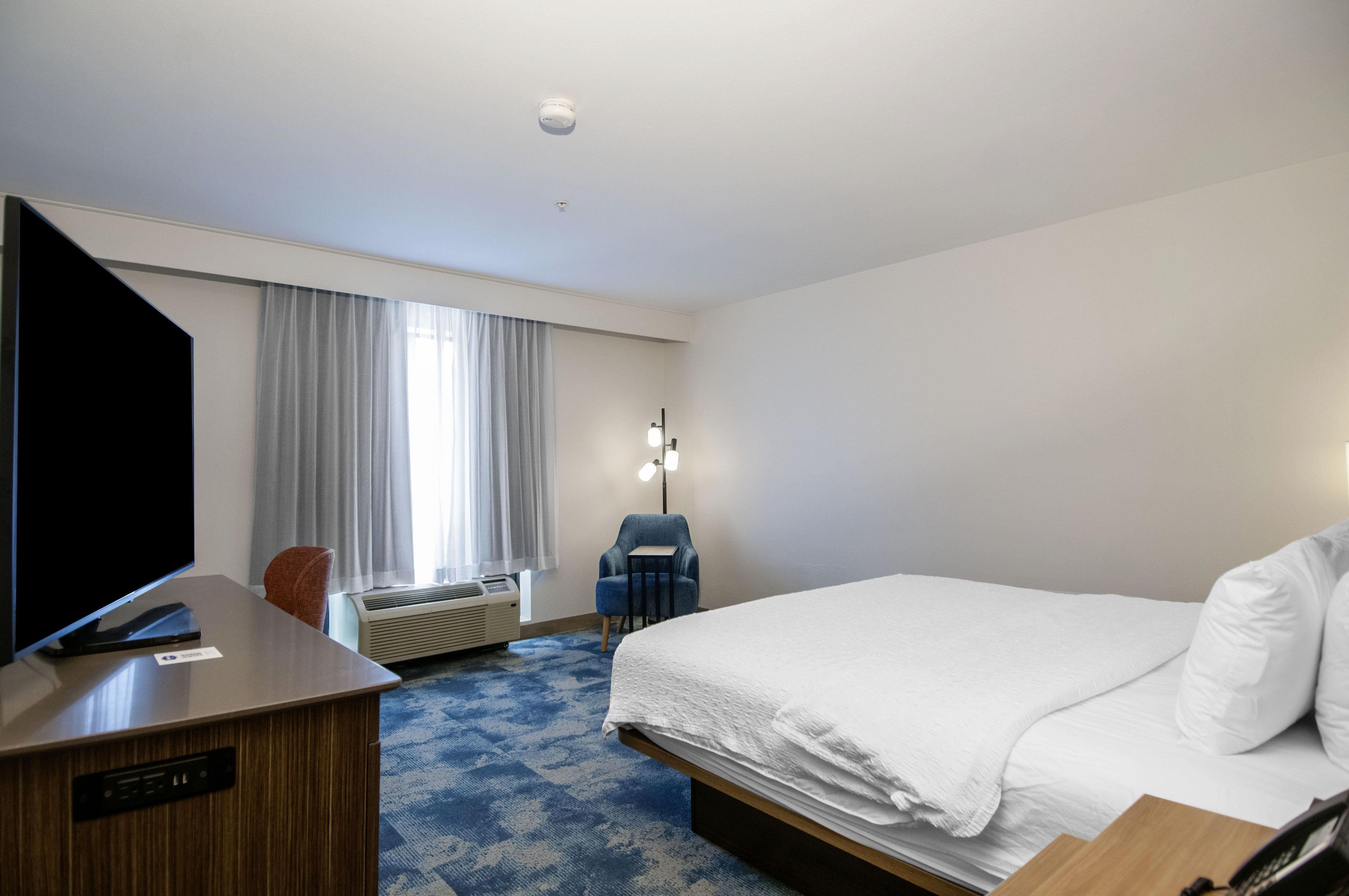 A hotel room with a bed, a desk, a chair, a window with curtains, and a wall-mounted lamp.