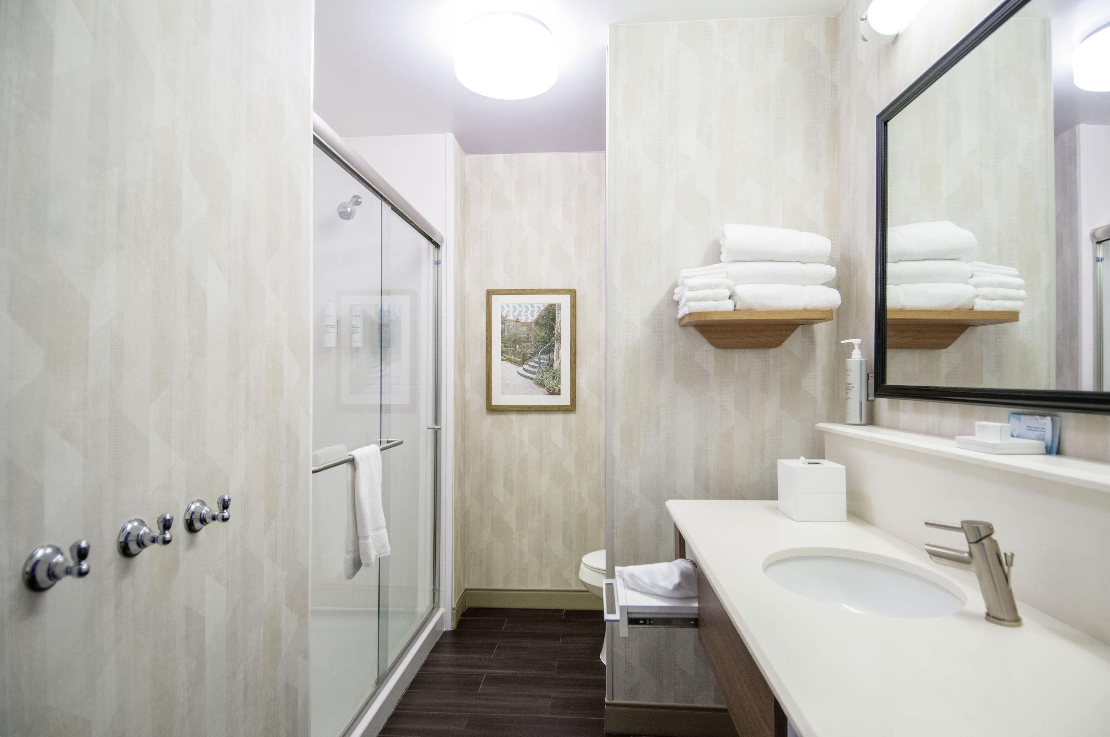 Un bany modern amb porta de dutxa de vidre, un lavabo blanc, un mirall i una fotografia encadrada a la paret.