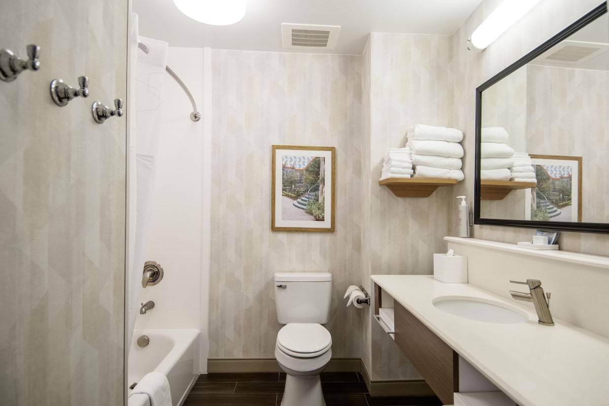 Un baño con bañera, inodoro, lavamanos y espejo. Hay toallas apiladas en un estante y un cuadro en la pared.