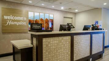 A reception desk with a 'Welcome to Hampton' sign, a 'Hilton' logo, and a digital kiosk.
