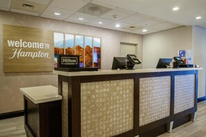 A reception desk with a 'Welcome to Hampton' sign, a 'Hilton' logo, and a digital kiosk.