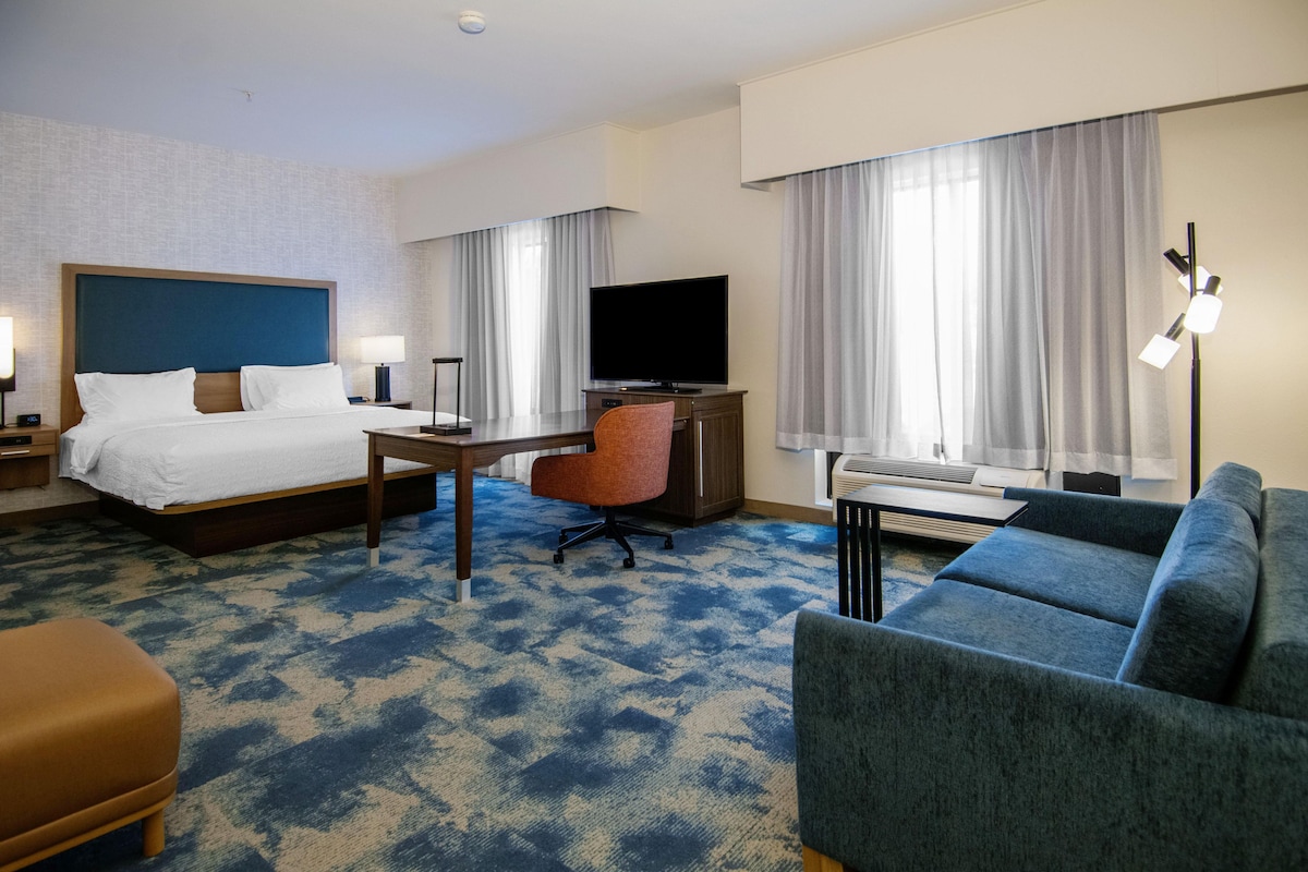 Habitación de hotel con cama, escritorio, televisor, sofá y un amplio ventanal con cortinas.