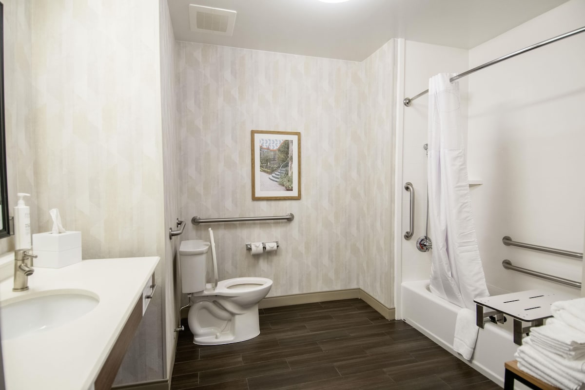 Un baño con inodoro, bidé y bañera. Hay un pasamanos y un cuadro en la pared.