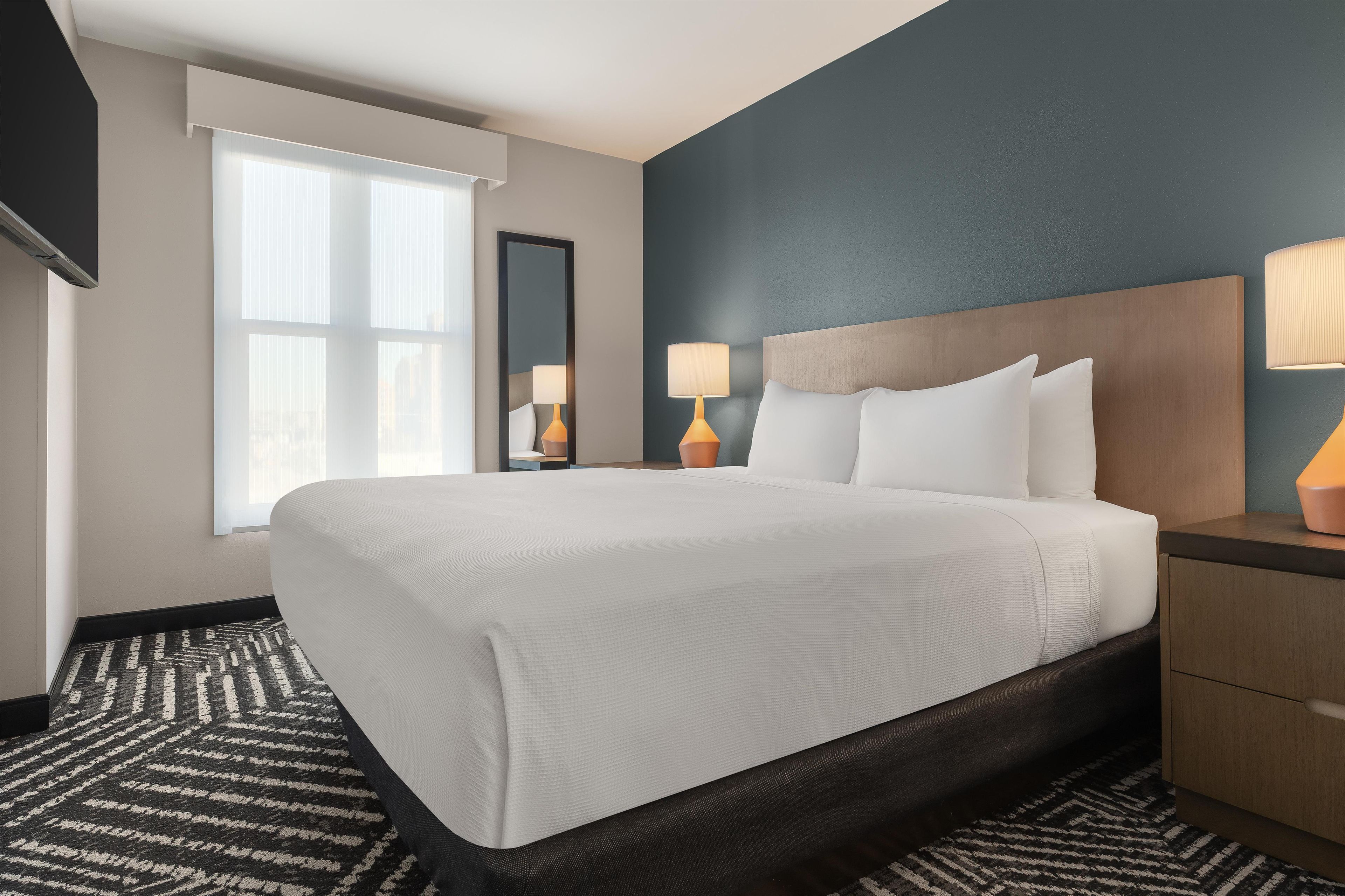 Une chambre d’hôtel avec un grand lit, une table de chevet, un miroir et une fenêtre avec des stores.