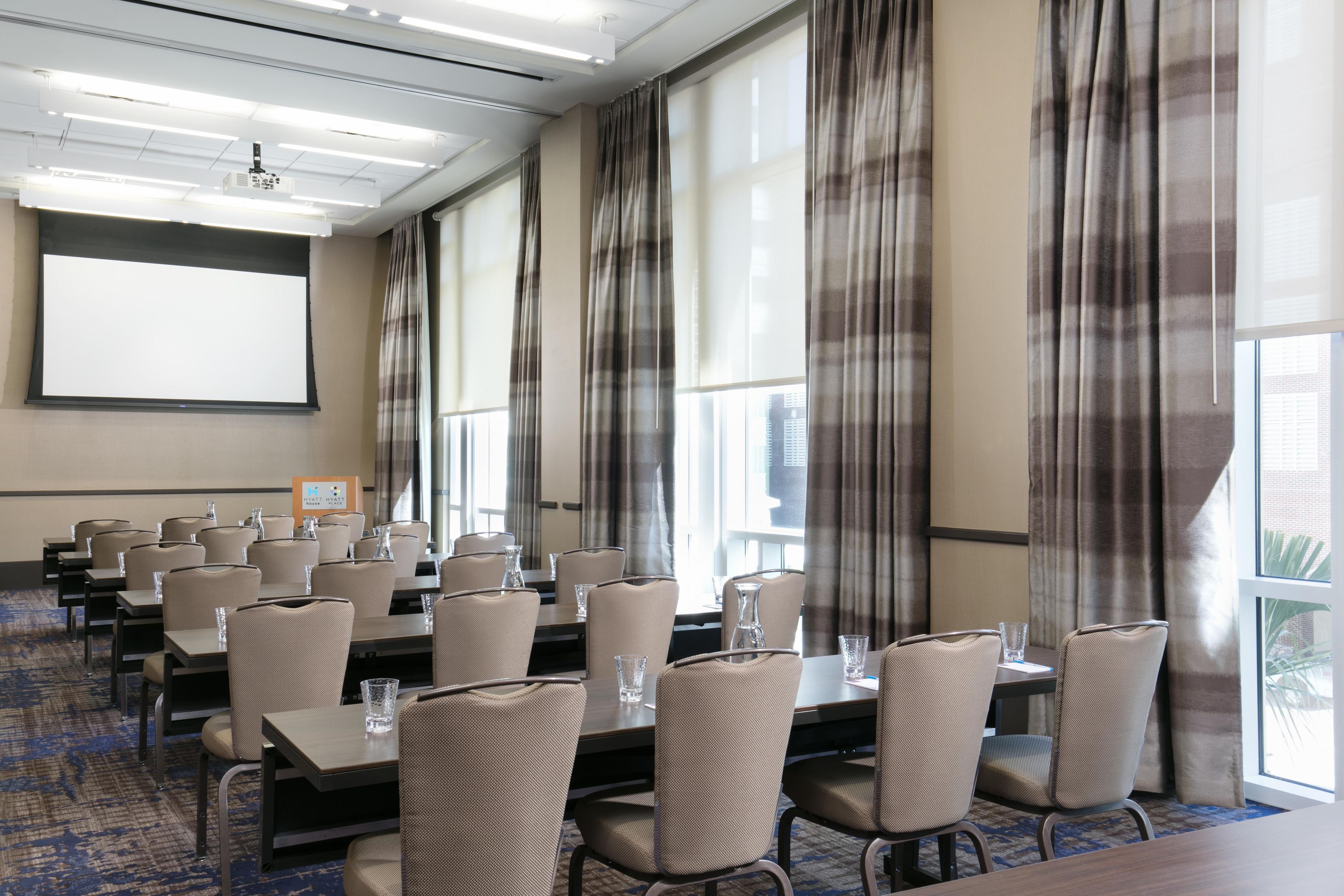 Une salle de conférence équipée d’un écran de projection, de grandes fenêtres avec des rideaux, ainsi que de rangées de tables et de chaises.