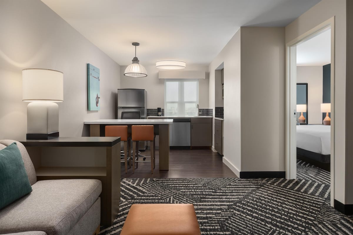 Ett modernt hotellrum med en soffa, ett litet bord, en köksdel med kylskåp och ett stort fönster.