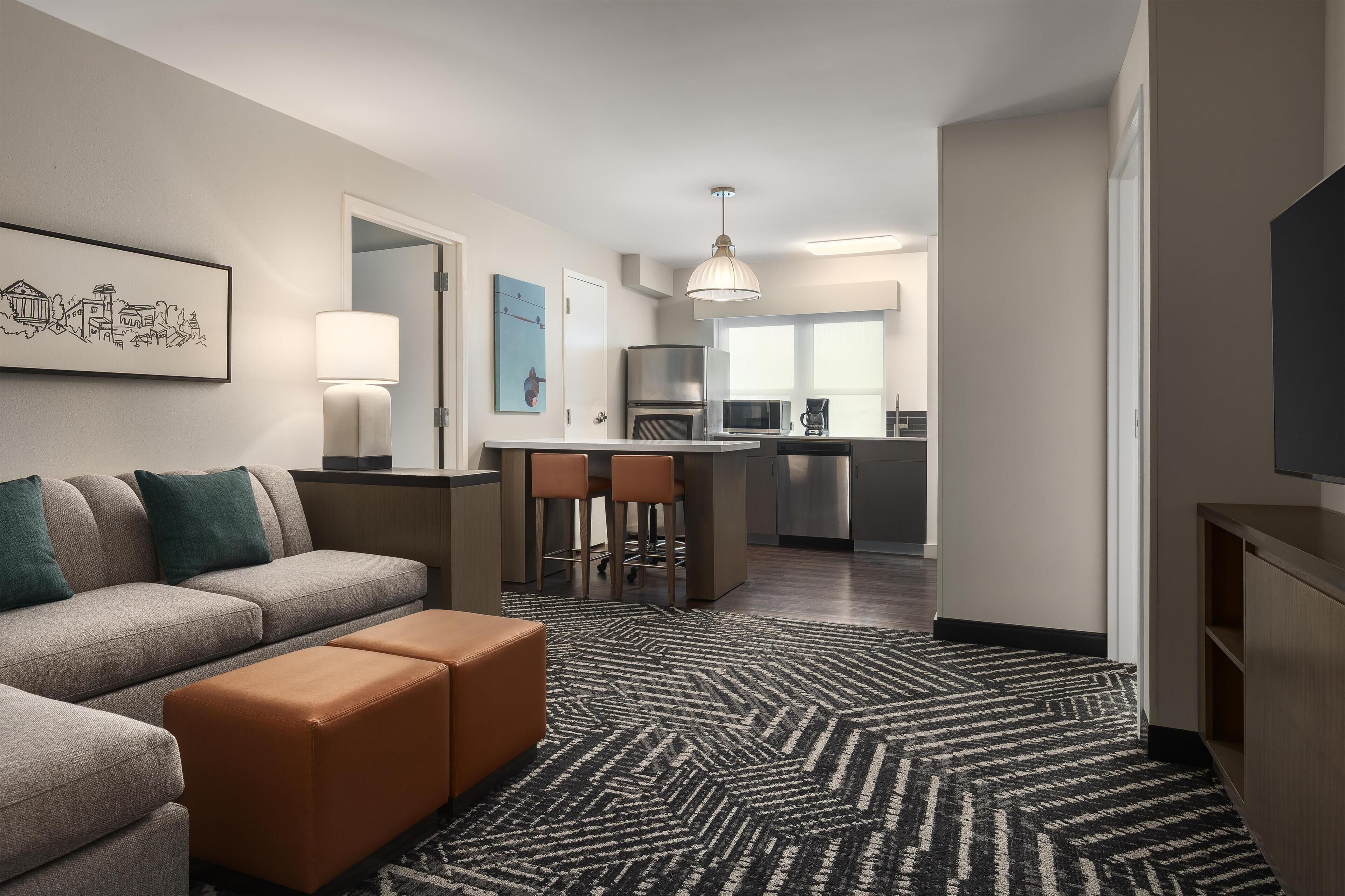 A modern hotel room with a sofa, ottoman, kitchen area, and a television.