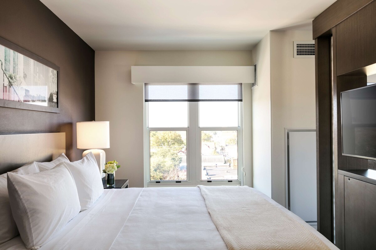Ein Hotelzimmer mit Bett, Fernseher, Fenster mit Blick und einem Wandbild.