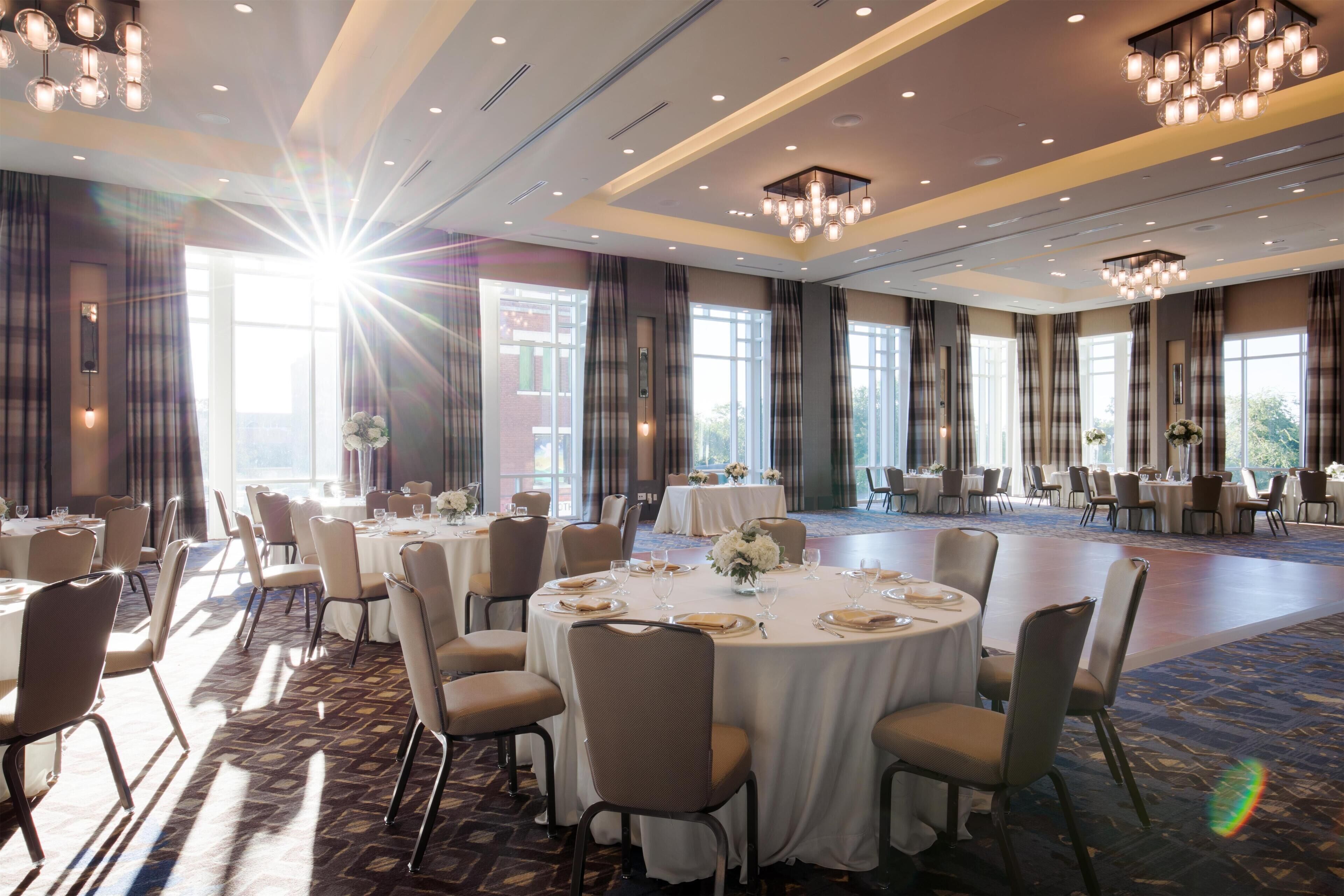 A banquet hall with round tables set for dining, large windows, and chandeliers.