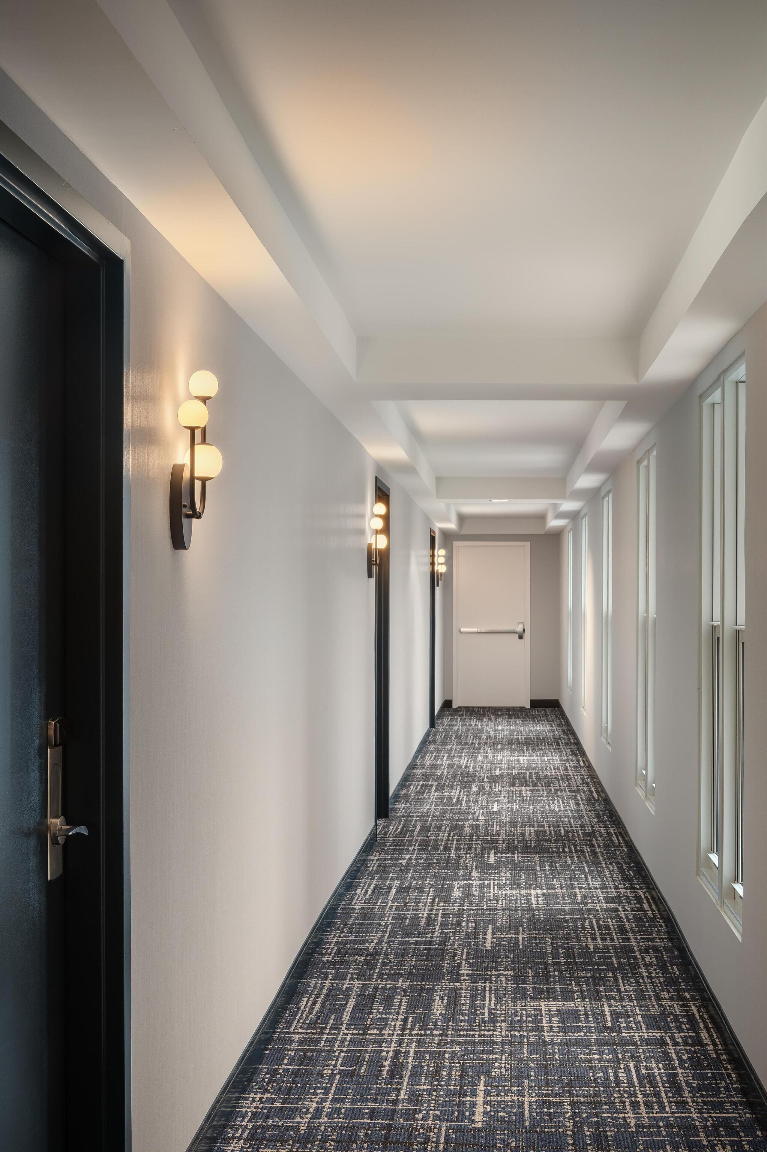 Un pasillo con alfombra estampada, luminarias fijadas a la pared, una puerta al final y ventanas a la derecha.