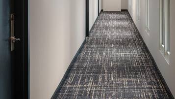 Un pasillo con alfombra estampada, luminarias fijadas a la pared, una puerta al final y ventanas a la derecha.