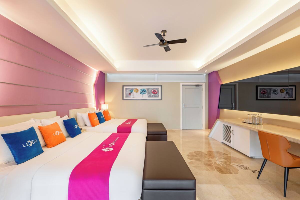 A hotel room with a modern design, featuring a large bed with a pink and white bedspread, multiple pillows, a desk with a chair, a ceiling fan, and artwork on the walls.