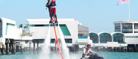 A jet ski with a person standing on its back, another person seated, and a building with a flag in the background.