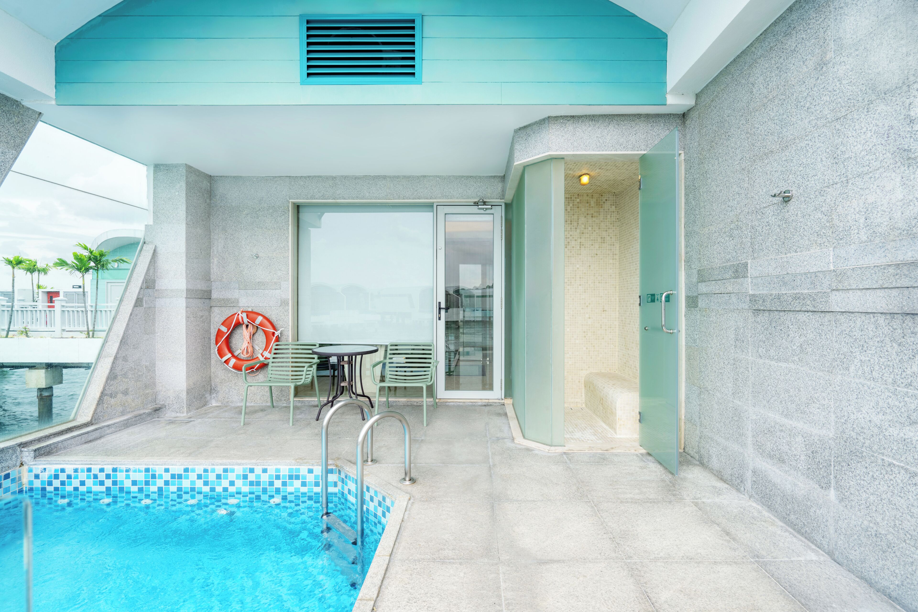 A modern indoor pool area with a clear glass wall, a lifebuoy, and a seating area with a table and chairs.