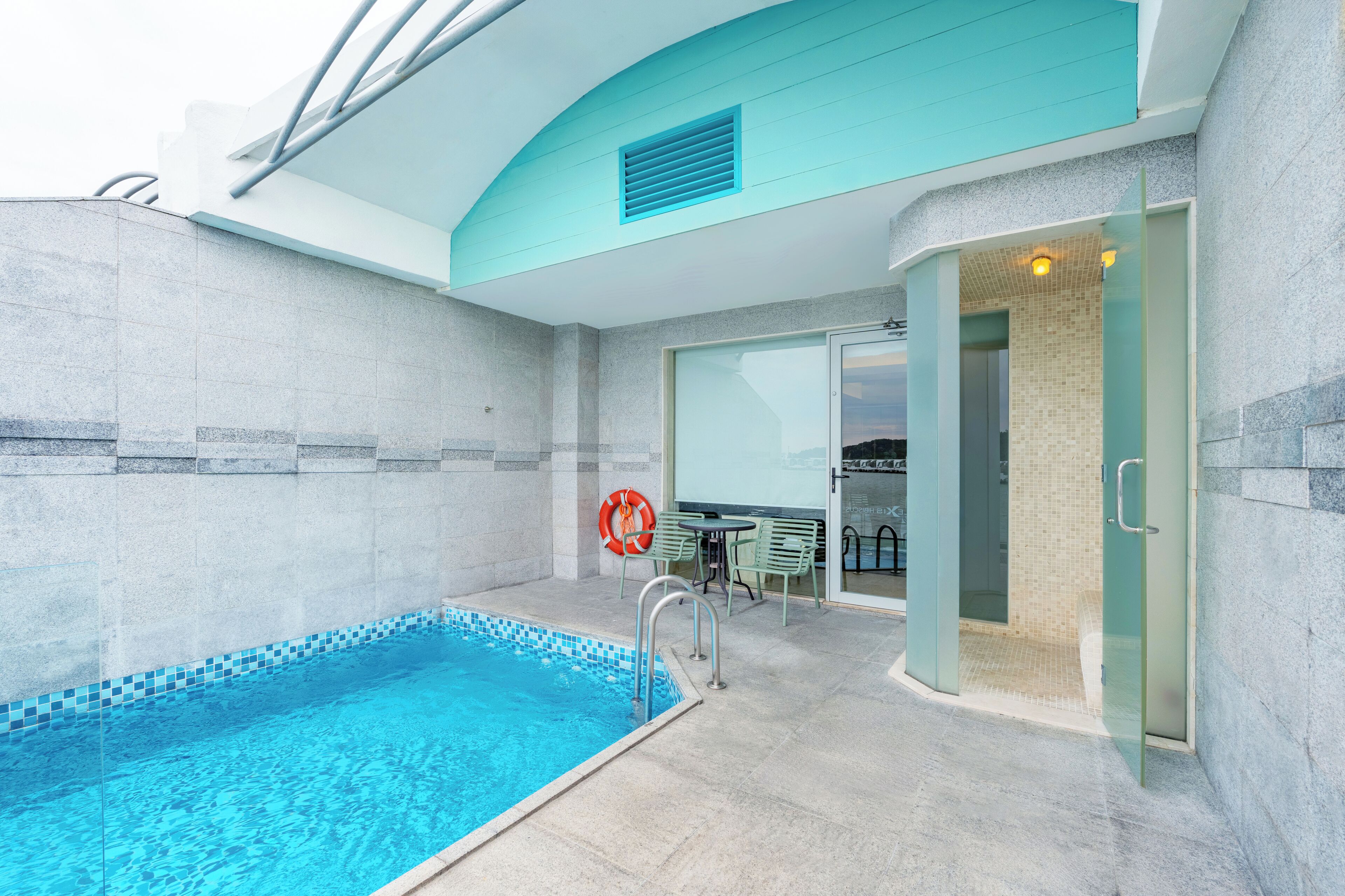 En swimmingpool med fliset kant, en redningskrans og en glasdør, der fører til et overdækket område med et bord og stole.