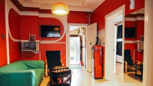 A room with a green sofa, a small table with a chessboard, a vintage red refrigerator, and a television mounted on the wall.