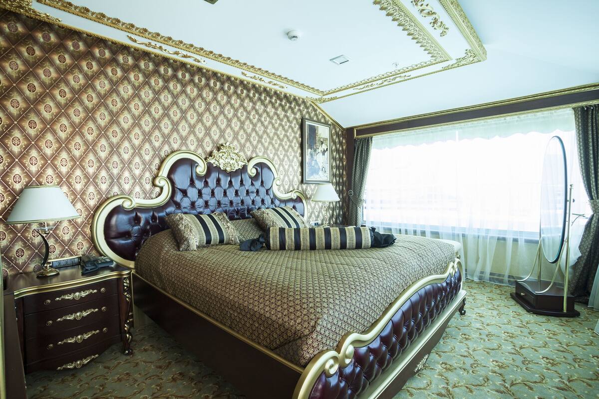 A large bed with a tufted headboard, a nightstand with drawers, a lamp, and a large window with curtains.
