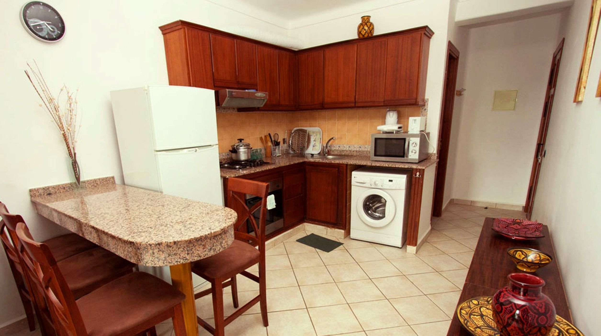 A compact kitchen with wooden cabinets, a white refrigerator, a microwave, a washing machine, and a granite countertop.