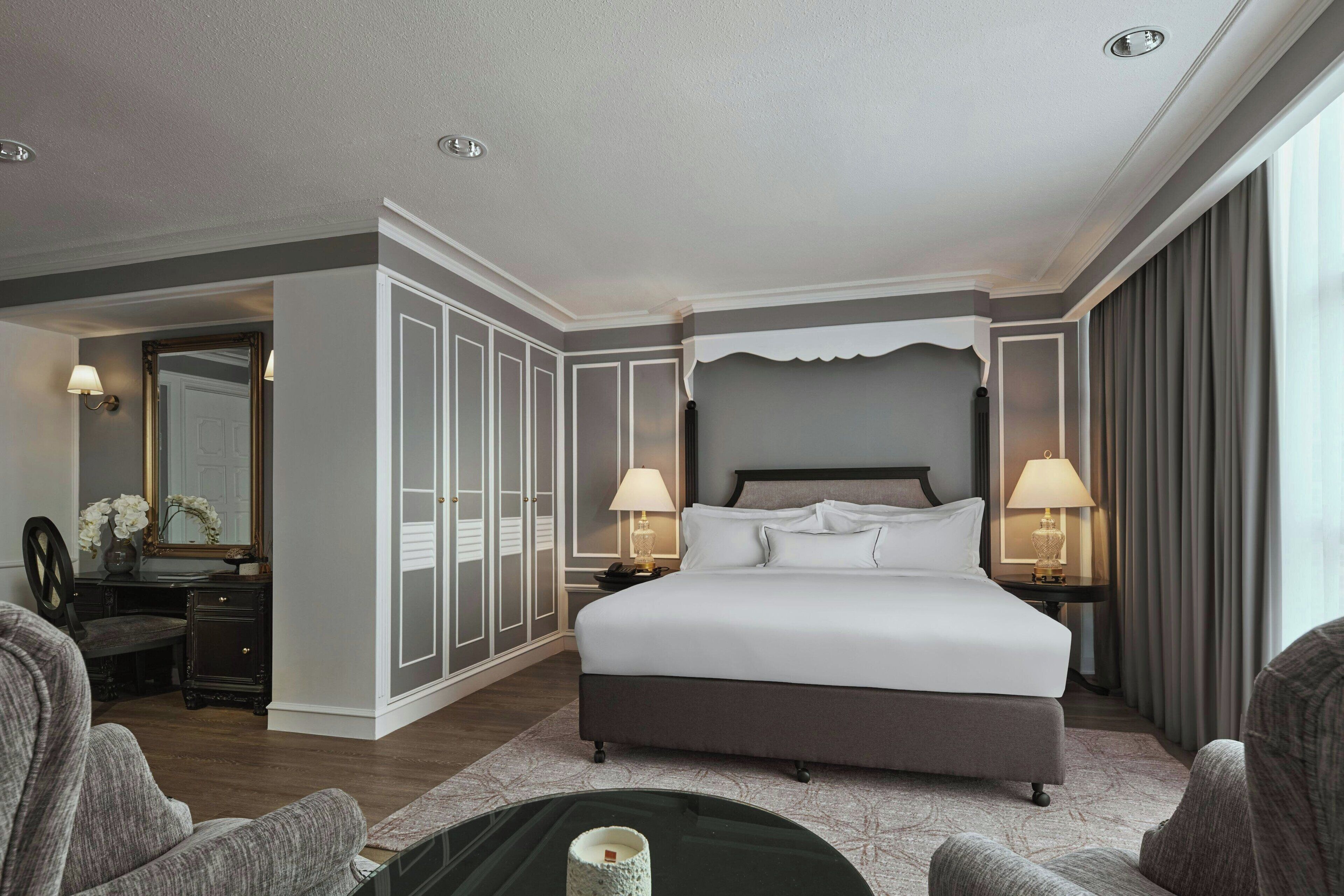 A spacious hotel room with a large bed, two bedside tables with lamps, a wardrobe, and a sitting area with a coffee table.