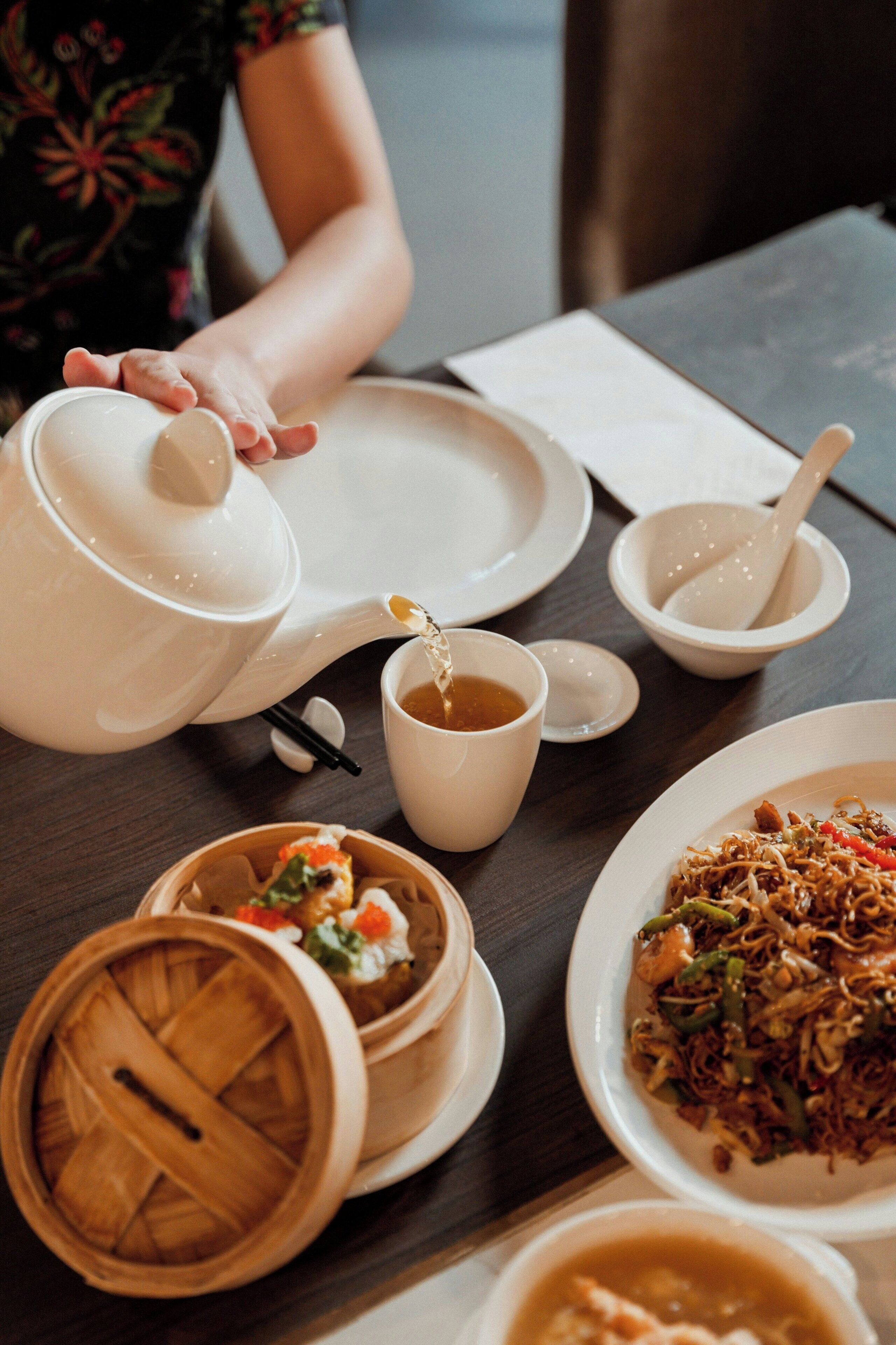 Een eettafel met een theepot, kopjes en diverse gerechten, waaronder een kom noodles en een kom soep.