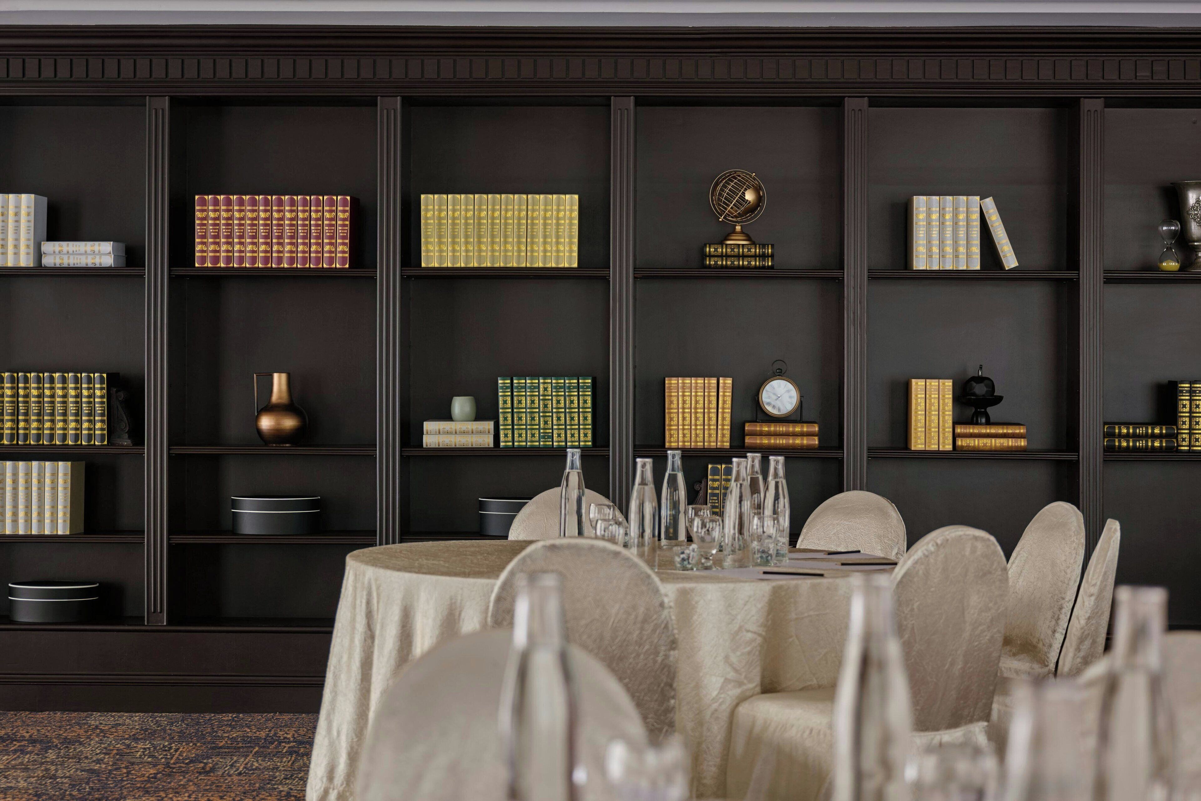 A dining area with a round table set for six, surrounded by upholstered chairs. Behind the table, there is a bookshelf with various books and decorative items.