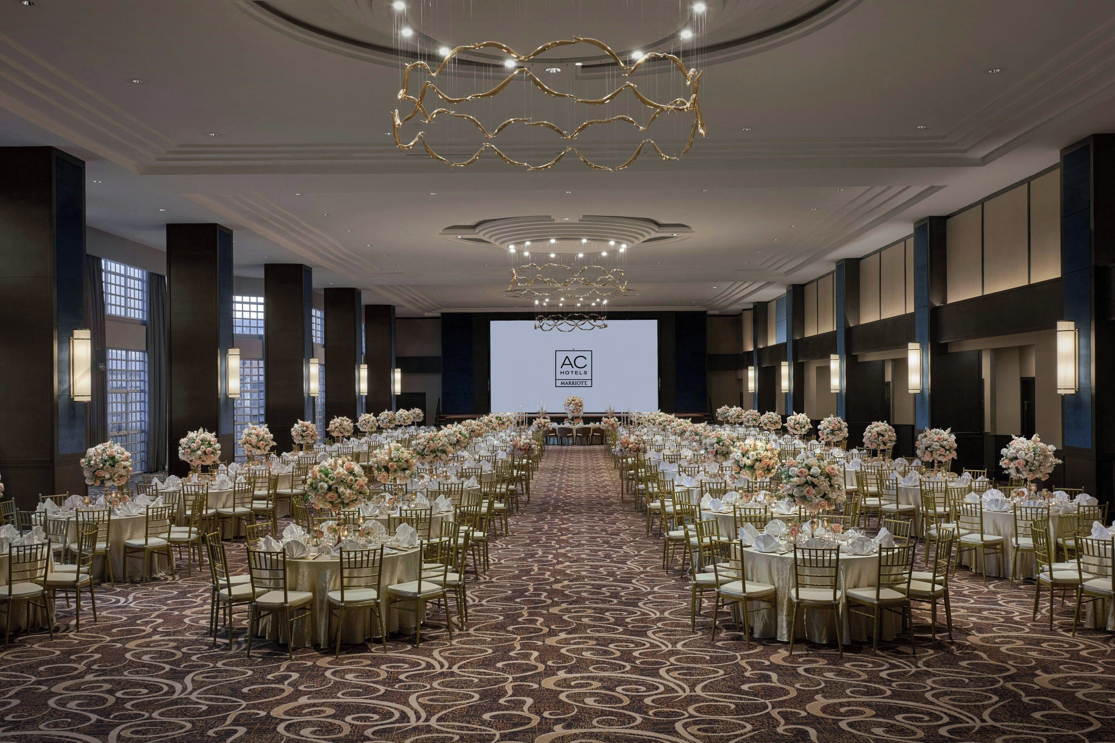 Un amplio salón de conferencias con filas de mesas redondas, cada una con sillas doradas y manteles blancos, decorado con arreglos florales.