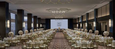 Une grande salle de conférence, avec de longues rangées de tables rondes, chacune agrémentée de chaises dorées et de nappes blanches, et décorée de compositions florales.