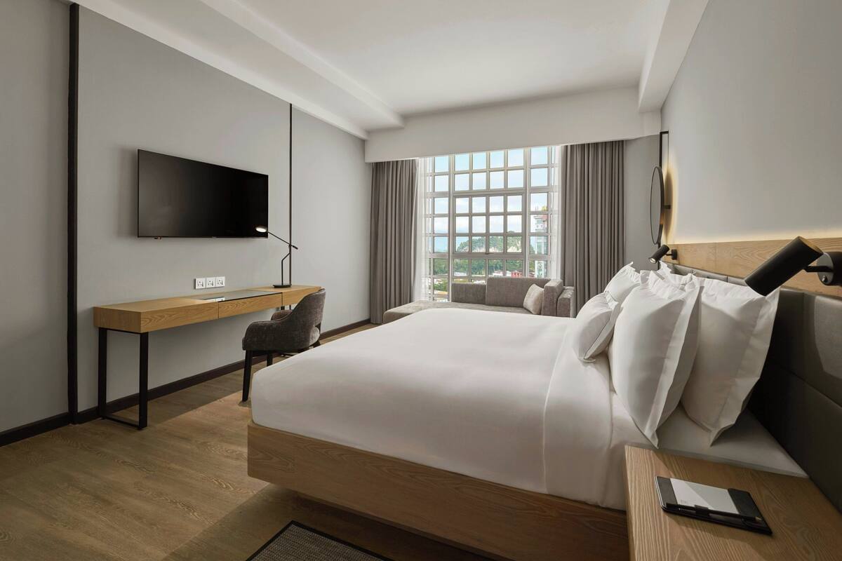 Ein modernes Hotelzimmer mit einem großen Bett, einem Schreibtisch, einem Flachbildfernseher und Blick auf die Stadt durch große Fenster.