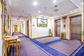Un hall d’hôtel avec un long couloir, un ascenseur, un comptoir d’accueil et un tableau encadré accroché au mur.