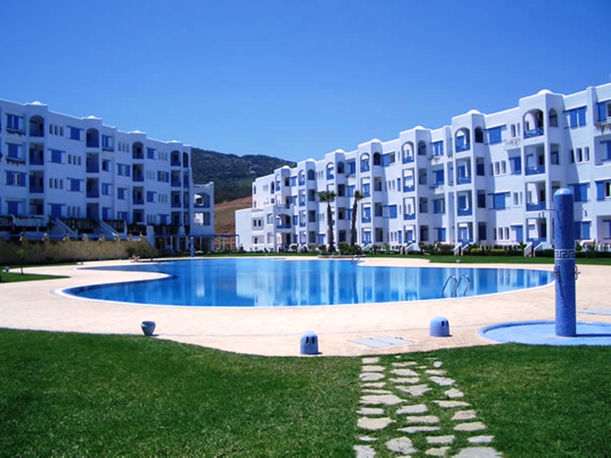 Un hotel moderno con una piscina central, rodeado de céspedes cuidados y un cielo azul despejado.