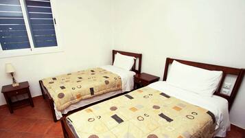 Una habitació amb dos llits individuals, una tauleta de nit amb una làmpada i una finestra amb persianes.