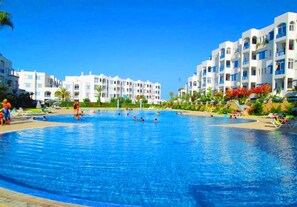 Una gran piscina rodeada de edificios blancos con balcones, palmeras y flores rojas.